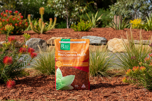 Bag of Red Cypress Mulch in a garden setting with plants and rocks.